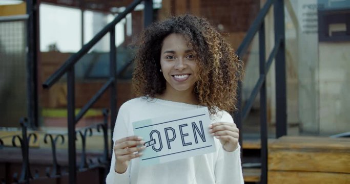 Beautiful African American black female standing with an open sign near her small cafe, smiling and looking into camera. Small business concept. 4K UHD 60 FPS SLOW MOTION
