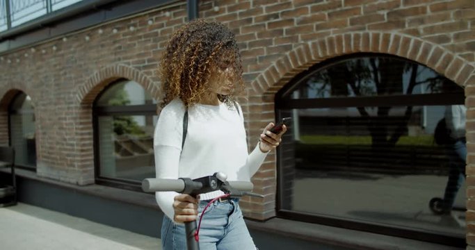 TRACKING attractive African American black female walking in the street with an electric scooter, checking her phone. 4K UHD 60 FPS SLOW MOTION