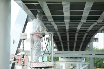 Professional worker in a protective suit and mask paints a bridge from the spray gun  stand on the hydraulic lift.