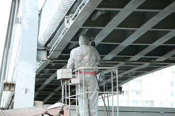 Professional worker in a protective suit and mask paints a bridge from the spray gun  stand on the hydraulic lift.