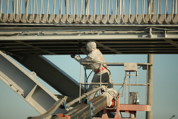 Professional worker in a protective suit and mask paints a bridge from the spray gun  stand on the hydraulic lift.