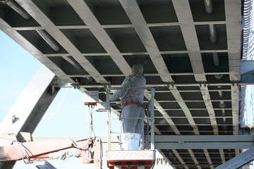 Professional worker in a protective suit and mask paints a bridge from the spray gun  stand on the hydraulic lift.