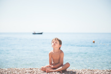 Fototapeta na wymiar beautiful relaxing little caucasian kid sitting on summer sea beach with closed eyes dreaming during sunbathe yoga training