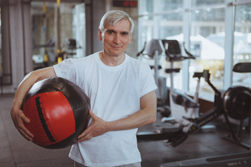Cheerful senior man smiling to the camera, holding medicine ball at the gym. Happy healthy elderly sportsman enjoying working out at sport studio