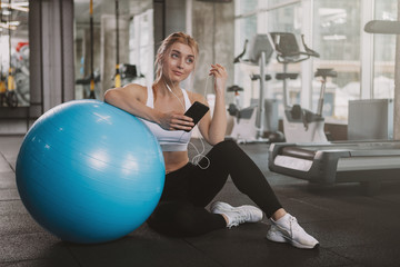 Gorgeous happy healthy young sportswoman using her smart phone at the gym, copy space. Lovely blond haired athletic woman relaxing after working out with fitness ball, copy space