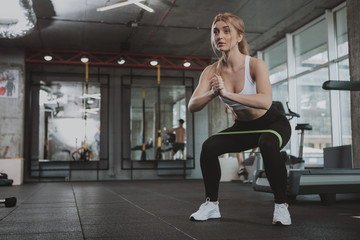 Fototapeta na wymiar Full length shot of a beautiful blond sportswoman doing squats with rubber resistance bans, copy space. Attractive healthy sportswoman working out at the gym, squatting with resistance band