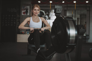 Attractive fitness woman looking concentrated, exercising on rowing machine at the gym, copy space. Healthy sportswoman working out at sport studio, using rowing machine