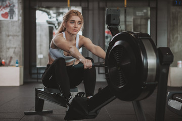 Fototapeta na wymiar Beautiful blond haired sportswoman doing cardio workout on row machine at the gym, copy space. Fitness woman using rowing machine at sport studio. Motivation, exercising concept