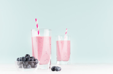 Blueberry milkshakes in transparent glass with blueberries in bowl, striped straw on white wood table and mint color wall, copy space.