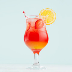 Fresh alcohol sunrise cocktail - red and yellow liquor, orange, ice, straw, strawberry in  wet glass on elegant pastel blue background.