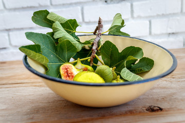 Fresh green fig tree leaf and ripe yellow and halved figs in an antique enamel bowl on a raw wood surface. 