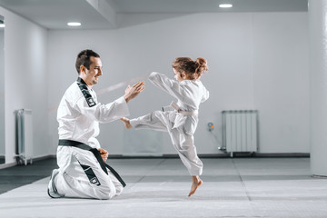 Smiling Caucasian taekwondo trainer in dobok kneeling and holding hand up while little girl kicking...