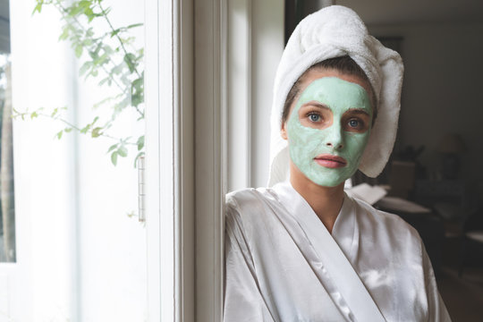 Woman in bathrobe wearing facial mask and leaning against the window