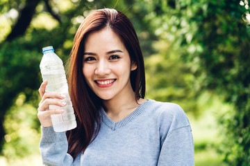 Beautiful asia woman drinking water from a bottle while relaxing and feeling fresh on green natural background at summer green park. Healthy lifestyle concept