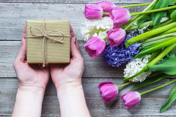 Female hands holding a gift or present box, flowers tulips, peony, hyacinth on a wooden table background. Top view