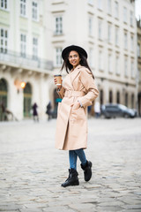 Naklejka premium Young woman wearing coat and black hat walking in the autumn city street and drinking take away coffee in paper cup. Breakfast to go.