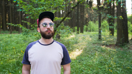 Naklejka na ściany i meble Young man with beard in sunglasses, hipster traveler in the sunny forest. Adventure, vacation, wanderlust, nature.