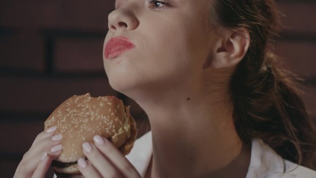 Hungry woman eating burger on brick wall background in fast food cafe