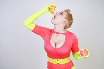 alternative caucasian girl with short hair dressed in sporty red spandex bodysuit and bright tights with green neon gloves. curvaceous woman holds a sliced grapefruit on a white studio background - obrazy, fototapety, plakaty