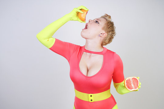 alternative caucasian girl with short hair dressed in sporty red spandex bodysuit and bright tights with green neon gloves. curvaceous woman holds a sliced grapefruit on a white studio background