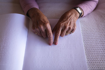 Blind active senior woman hands reading a braille book on bed in bedroom at home