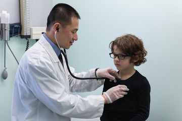 Young asian male doctor examining caucasian boy patient with stethoscope in clinic 