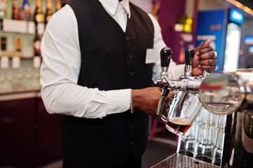 Close up hands of african american bartender at bar pouring from tap fresh beer into the glass in pub.
