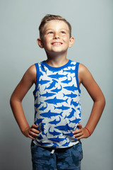 Cute little boy in white and blue sleeveless shirt posing in front of gray background. Portrait of fashionable male child. Smiling boy posing, looking up. Concept of children style and fashion. Studio
