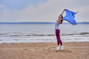 Fototapeta na wymiar Young slim brunette woman in sportswear doing sport dancing with a blue scarf. A woman is engaged on the sandy shore of a large river. Cloudy summer morning.