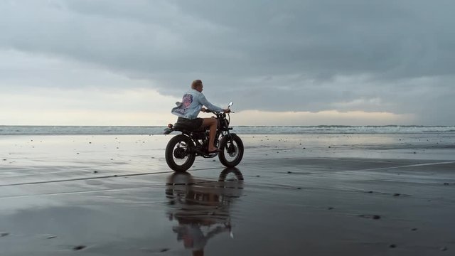 Young handsome hipster man riding modern custom motorcycle racer on the black sand beach near the water. Surfing spot with ocean waves.