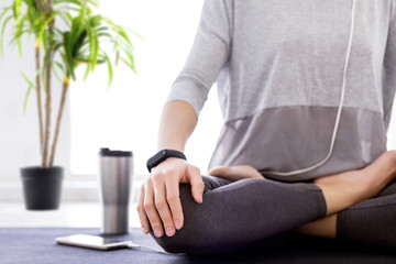 Fototapeta premium Closeup of slender female legs folded crossed in lotus position. Young athletic woman in grey clothes and fitness tracker is practicing yoga, meditating, relaxing while sitting in padma asana at home.