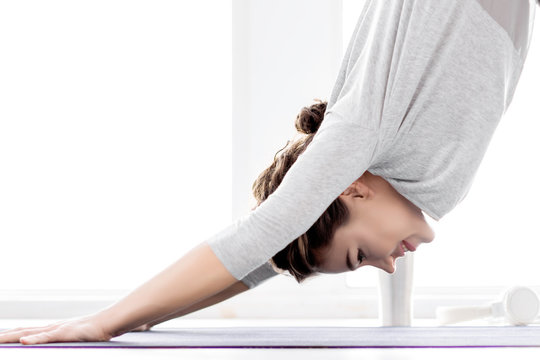 Portrait of smiling brunette female bent over to position downward facing dog. Young athletic woman in grey clothes is practicing yoga at light fitness studio. Healthy lifestyle concept.
