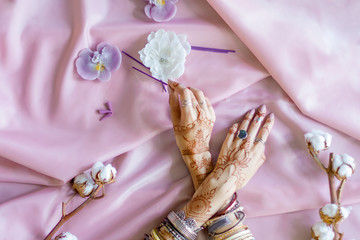 Female wrists painted with traditional Indian oriental mehndi ornaments. Hands dressed in bracelets and rings hold aroma stick. Pink fabric with folds, cotton branches and candles on background.