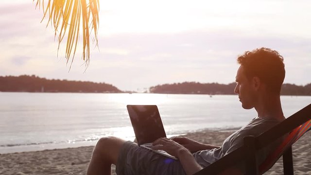 man working on laptop computer at sunset beach, banking online, freelancer job or telework