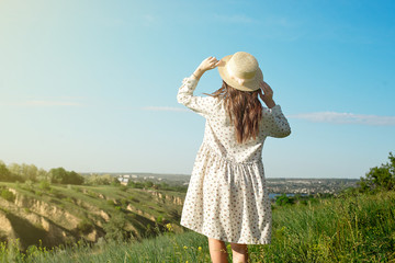 Satisfied brunette in a long summer dress wearing a straw hat is standing in the tall grass with...