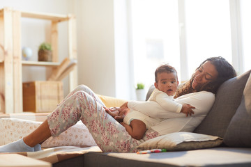 Smiling careful loving young African mother with wavy hair sitting on comfortable sofa and hugging baby while they relaxing together - obrazy, fototapety, plakaty