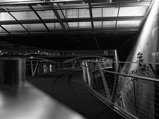 Perspective view of pedestrian overpass above the River Thames with steel fence and guard rails at night. Black and white image. Bridges and the river are seen in the background.