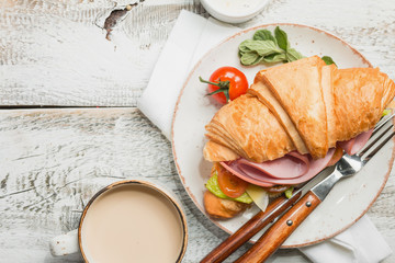 Fresh croissant sandwich with ham, cheese and salad leaf with coffee on white table, top view