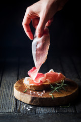 Female hand putting prosciutto on bread over dark wooden background. Sandwich cooking. - obrazy, fototapety, plakaty