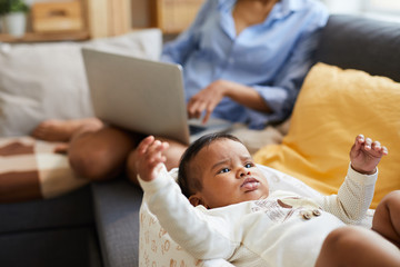 Close-up of angry frowning cute black baby boy in bodysuit lying in cocoon and gesturing hands while mother working in background