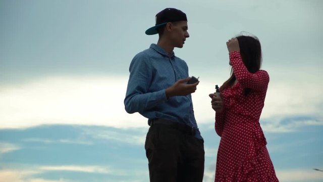 Handsome man vaping e-cigarette and talking with pretty woman smoking electronic tobacco heating system  on the cloudy sky background. Young people relaxing outdoors.