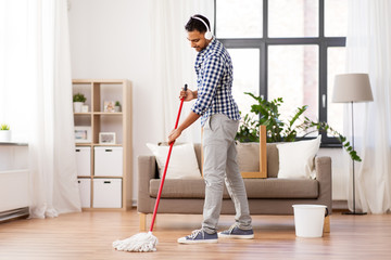 household, housework and people concept - happy indian man in headphones with mop and bucket...