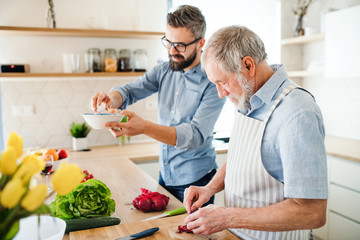 An adult hipster son and senior father indoors at home, cooking.