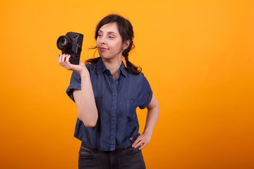 Fototapeta premium Portrait of young female photographer holding her camera in studio over yellow background