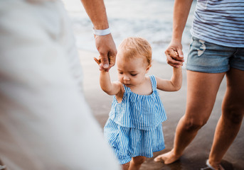A midsection of parents with toddler daughter walking on beach on summer holiday.