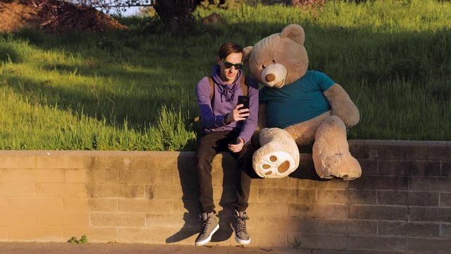 Caucasian guy consults teddy bear before taking a selfie together. Sitting on brick wall in shady area. Rustic scene. Slow motion.