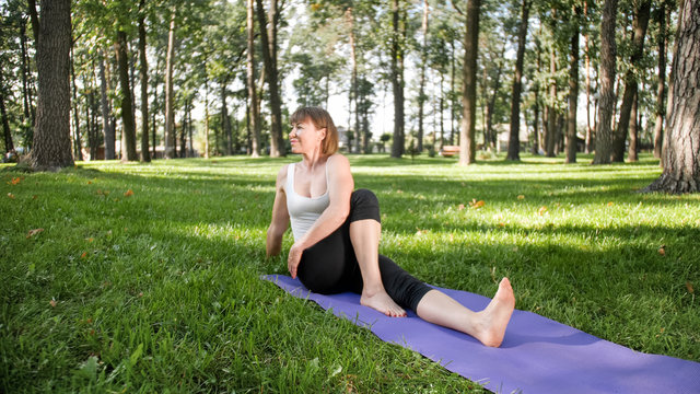 Image of smiling middle aged woman in fitness clothes doing stretching and yoga exercises. WOman meditating and doings sports on fitness mat on grass at park