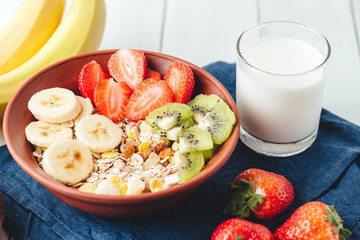 Cereal muesli with strawberries, banana and kiwi in the bowl, glass with milk on wooden white background, healthy breakfast