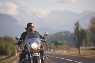 Portrait of handsome bearded motorcyclist in black leather jacket holding motorcycle handles on country roadside on blurred background of landscape, distant white mountain peaks.