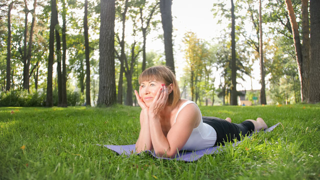 Image of mid aged smiling happy woman meditating and doing yoga exercises on grass at forest. Woman taking care of her physical and mental health while pracitising fitness and stretching at park